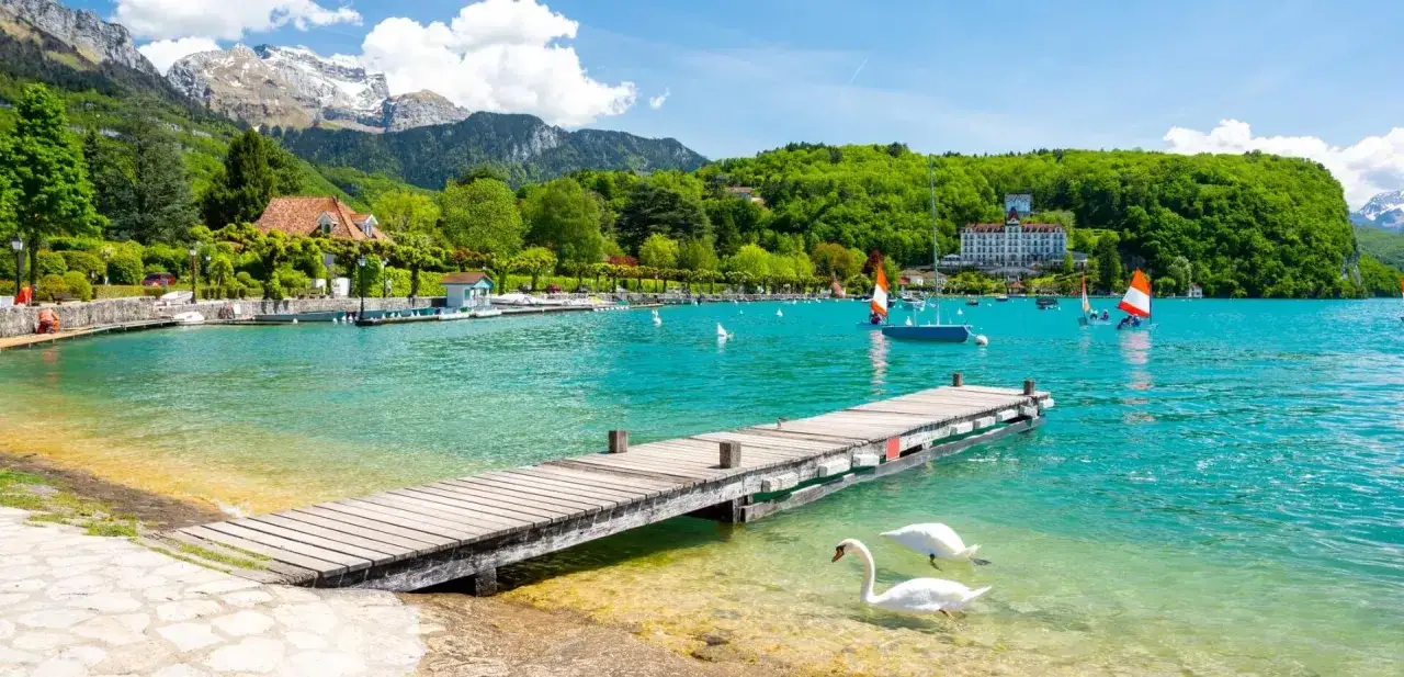 Deux cygnes nagent près d'un ponton en bois sur la plage de l'Impérial, avec des voiliers et un grand hôtel en arrière-plan.