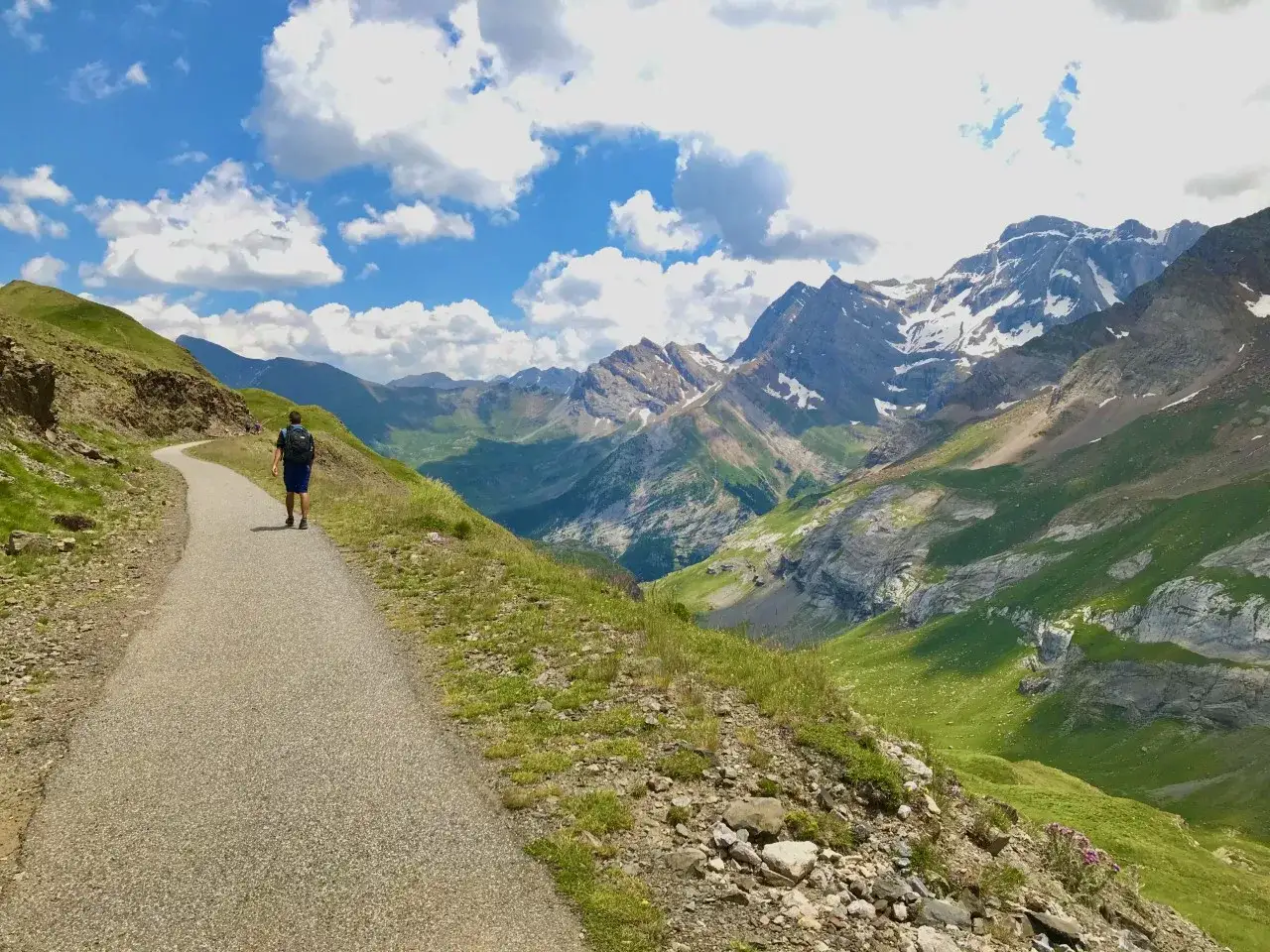 Un randonneur solitaire emprunte un sentier sinueux menant vers le col de tentes, dominant une vall&eacute;e verdoyante et des montagnes majestueuses.