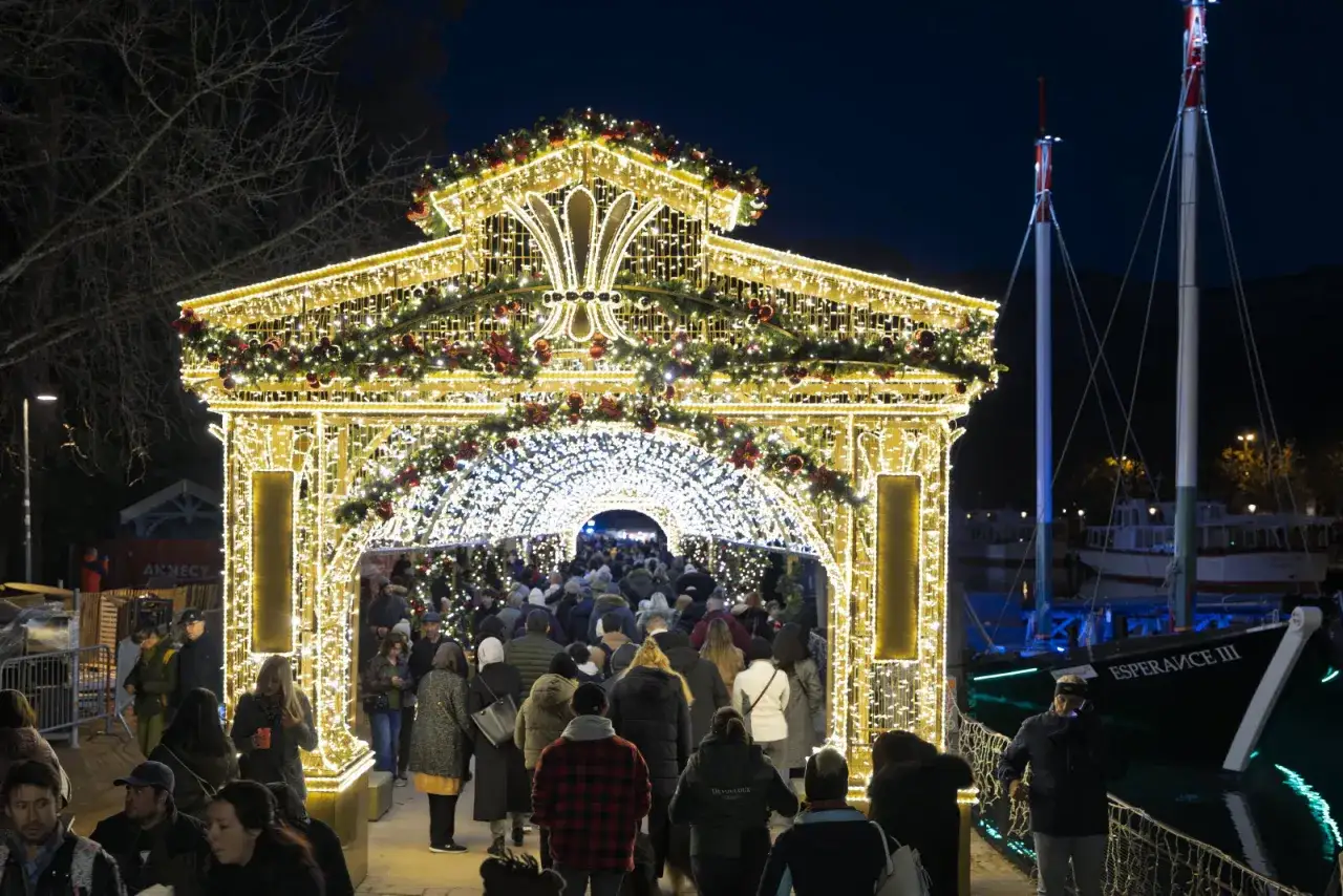 Une arche illumin&eacute;e de No&euml;l &agrave; Annecy en d&eacute;cembre, avec une foule traversant le tunnel de lumi&egrave;re. Des bateaux sont amarr&eacute;s sur le lac en arri&egrave;re-plan.