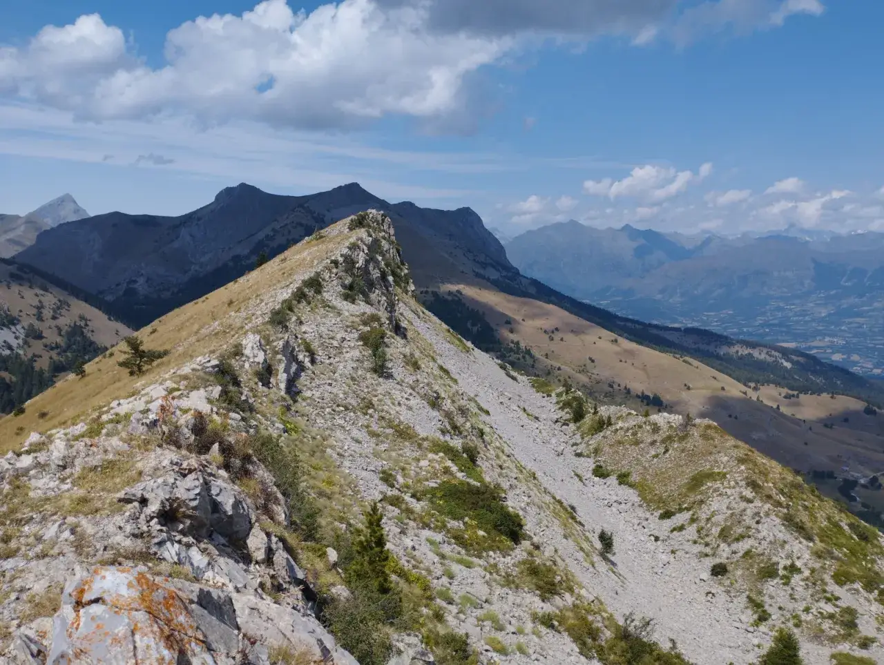 Vue panoramique depuis le col de Gleize, crête rocheuse et herbeuse dominant des vallées et des montagnes lointaines sous un ciel bleu et nuageux.
