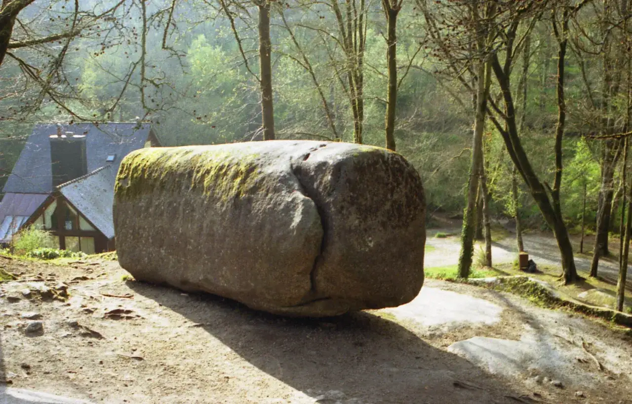 Un &eacute;norme rocher couvert de mousse, surnomm&eacute; la grotte du diable, repose sur un sentier bois&eacute; pr&egrave;s d'une maison.