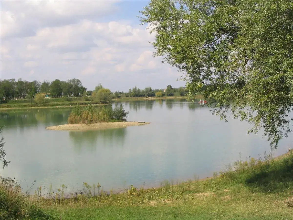 Le lac de Beaupuy, paisible, avec une petite &icirc;le verdoyante au centre. Un bateau rouge navigue doucement sur l'eau calme.