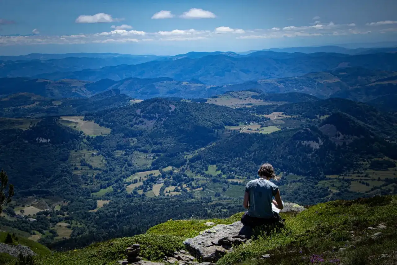 Vue panoramique depuis le pic du Lizieux. Une personne assise contemple la vallée verdoyante et les montagnes bleutées à l'horizon.