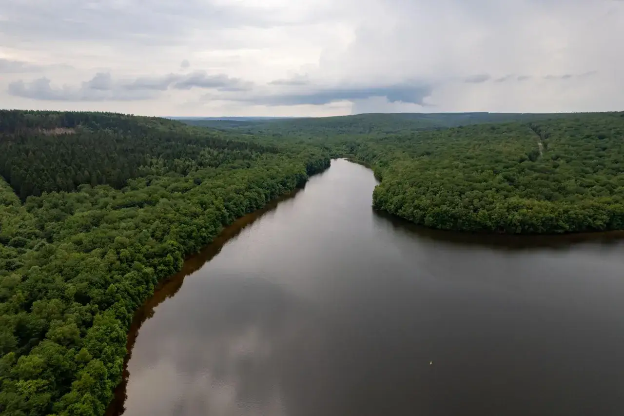 Vue aérienne du lac des Vieilles Forges, entouré de forêts denses sous un ciel nuageux. Une petite bouée orange flotte sur l'eau sombre.
