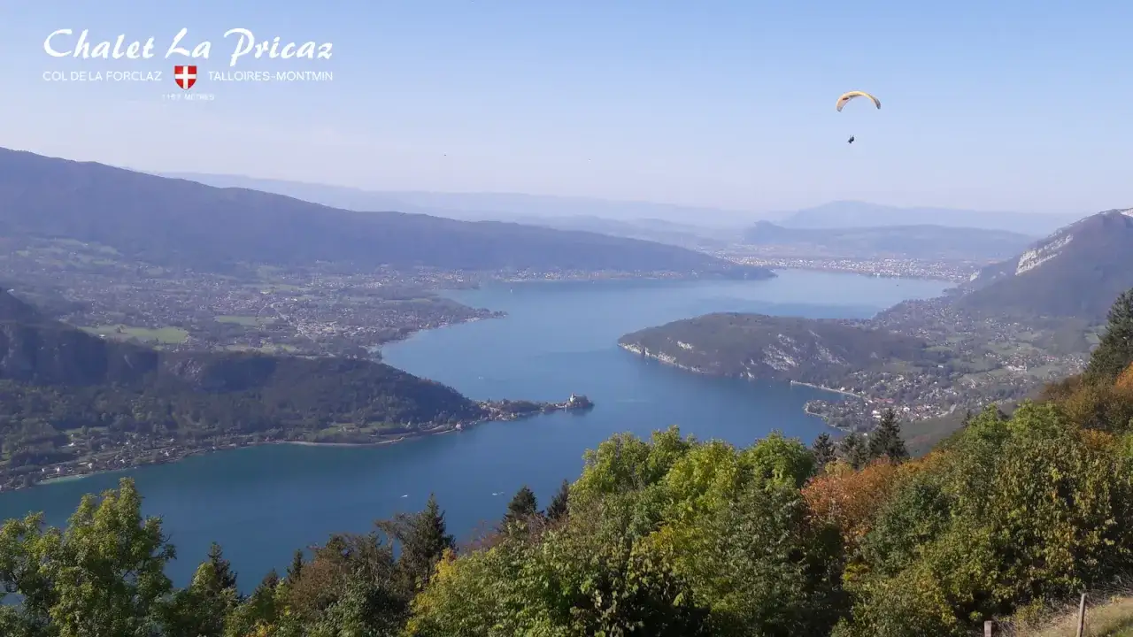 Vue panoramique du lac d'Annecy depuis le Col de la Forclaz. Un parapentiste survole le lac, offrant une perspective a&eacute;rienne unique.