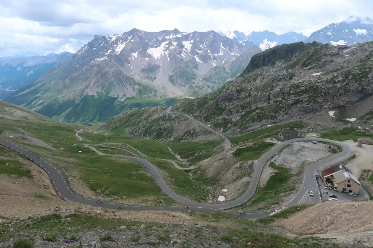 Vue spectaculaire du col du Galibier, avec ses lacets sinueux traversant des paysages alpins verdoyants et rocheux, sous un ciel nuageux.