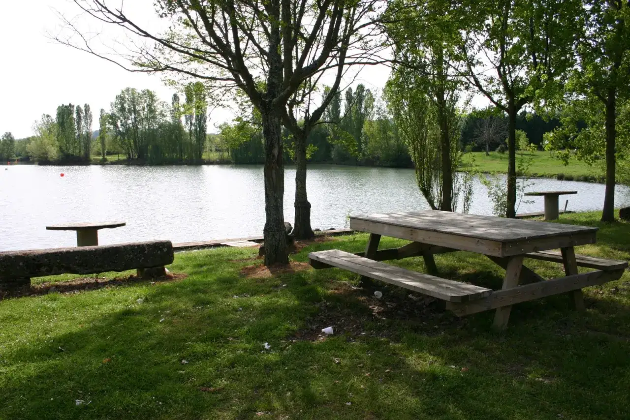 Aire de pique-nique au bord du lac de la Cadie, avec tables en bois et bancs en pierre, entourée d'arbres verdoyants.