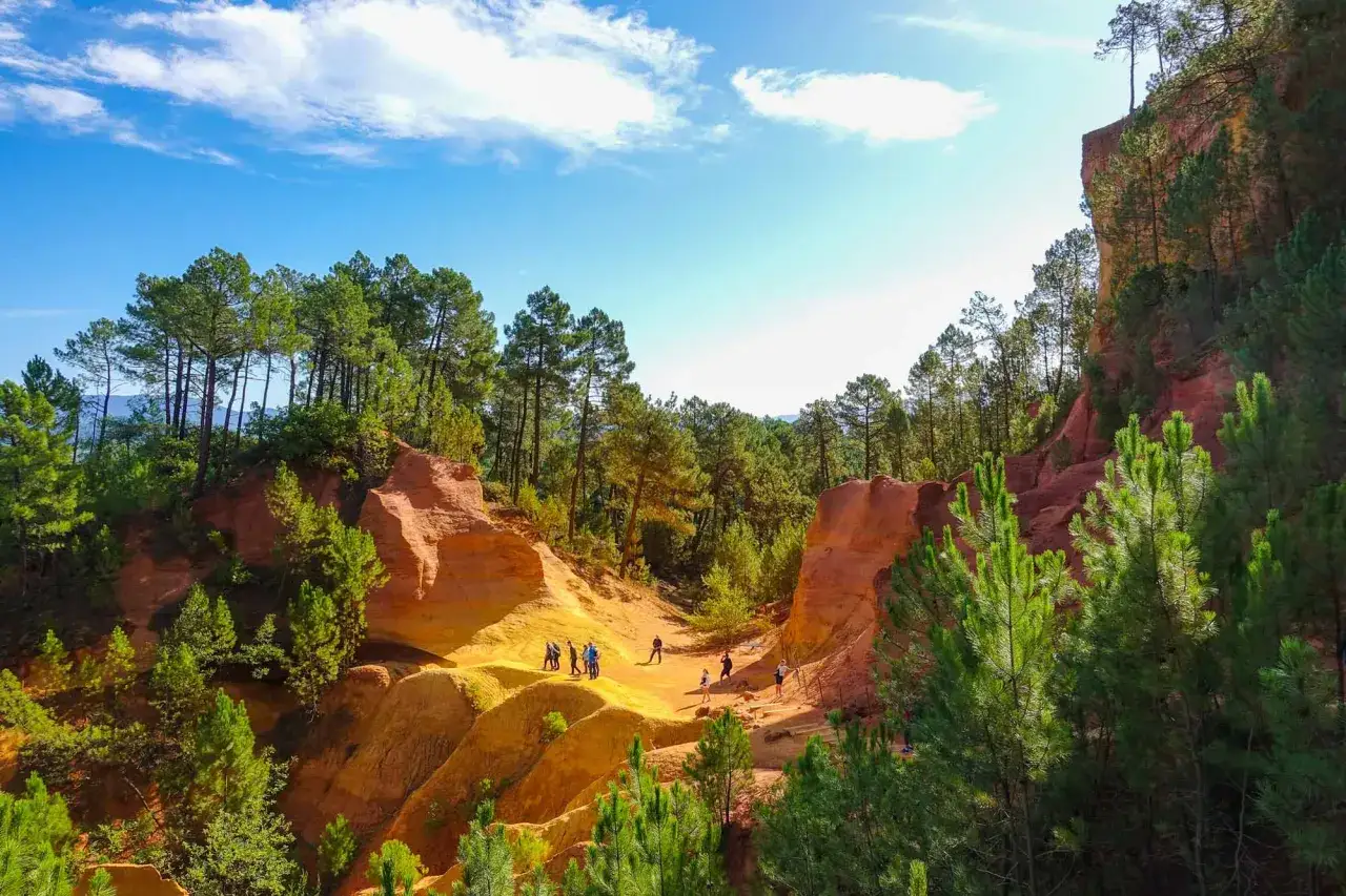 Des randonneurs explorent les paysages spectaculaires des ocres de Mormoiron, avec leurs falaises rouges et jaunes et leurs pins verts sous un ciel bleu.