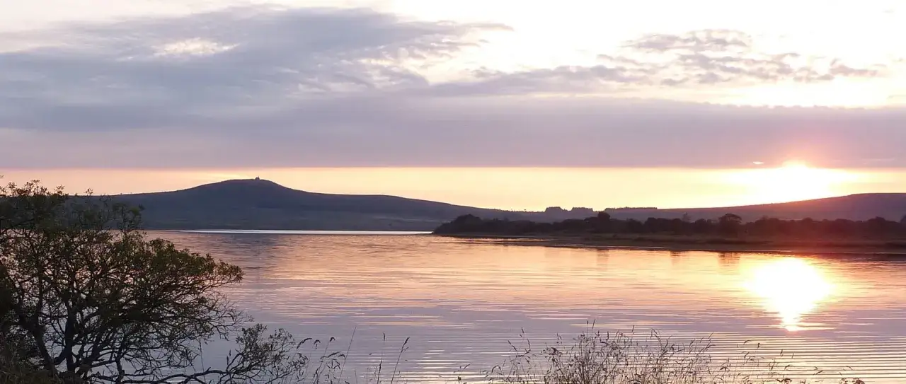 Coucher de soleil sur le lac de Brennilis, avec des collines et des arbres en silhouette.