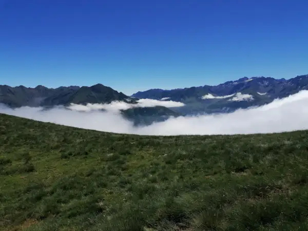 Vue panoramique depuis le col de Riou, avec des montagnes verdoyantes émergeant d'une mer de nuages sous un ciel bleu éclatant.