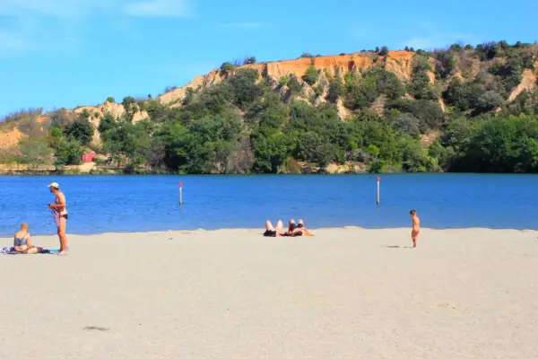 Plage de sable fin au lac de Vinça, avec des baigneurs profitant du soleil et de la vue sur la colline verdoyante.