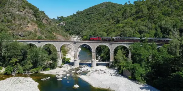 Le petit train des Cévennes traverse un viaduc de pierre au-dessus d'une rivière. La durée du trajet est une invitation à la découverte de ce paysage verdoyant.