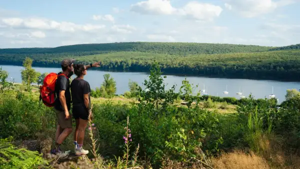Couple randonneurs admirant le lac des Vieilles Forges, avec des voiliers sur l'eau et une forêt dense en arrière-plan.