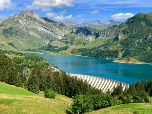 Vue panoramique du lac de Roselend, un barrage imposant bordé de forêts verdoyantes et de montagnes majestueuses sous un ciel bleu éclatant.