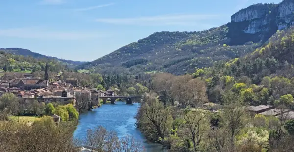 Vue panoramique de la gorge de l'Aveyron avec un village pittoresque, une rivière bleue et des collines verdoyantes sous un ciel clair.