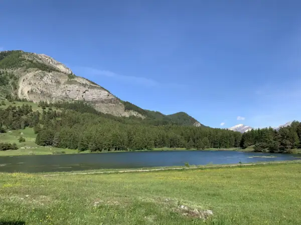 Le lac de Barbeyroux, paisible, reflète le ciel bleu. Des montagnes rocheuses et une forêt dense bordent ses rives verdoyantes.