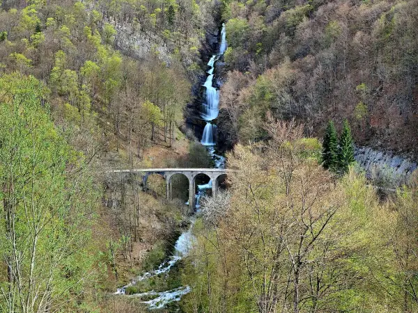La cascade du Bief de la Ruine dévale une falaise rocheuse, traversée par un pont en arc. La nature verdoyante environnante annonce le printemps.