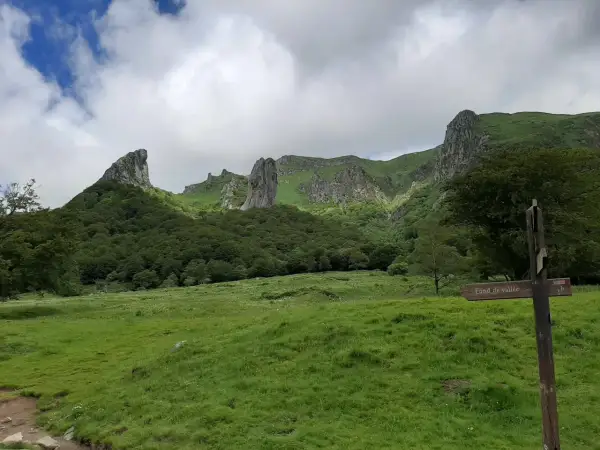 Paysage montagneux verdoyant avec des formations rocheuses spectaculaires. Un panneau indique "Fond de vallée", peut-être le chemin vers la cascade de la biche.