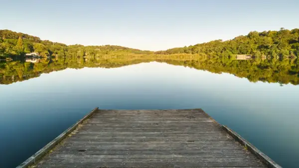 Un ponton en bois mène au lac Marion de Biarritz, reflétant les collines verdoyantes sous un ciel clair.