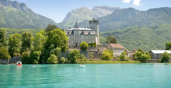 Un château majestueux surplombe un lac turquoise, invitant à aller à Annecy. Des montagnes verdoyantes en arrière-plan.