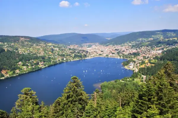 Vue panoramique du lac de Gérardmer, avec des voiliers sur l'eau bleue, entouré de collines verdoyantes et d'une ville pittoresque.