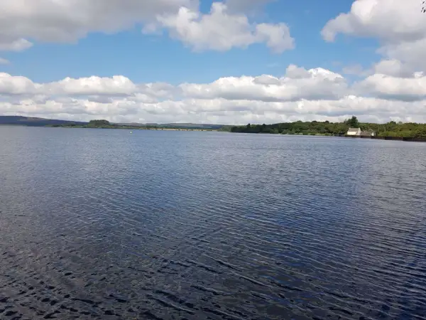 Vaste étendue d'eau calme sous un ciel bleu parsemé de nuages blancs. Le lac de Brennilis s'étend à perte de vue, bordé par une ligne d'arbres verdoyants et quelques constructions.