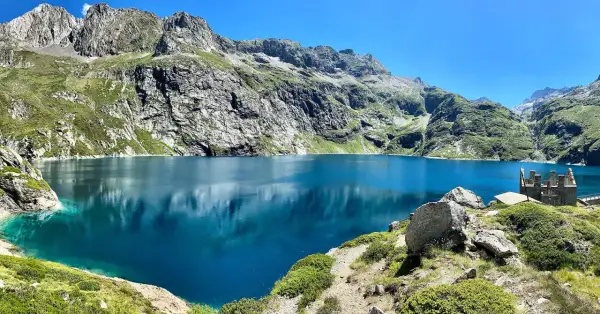 Le lac de Caillauas, d'un bleu profond, reflète les montagnes escarpées et verdoyantes sous un ciel clair. Une structure rocheuse rappelle un château sur la rive.