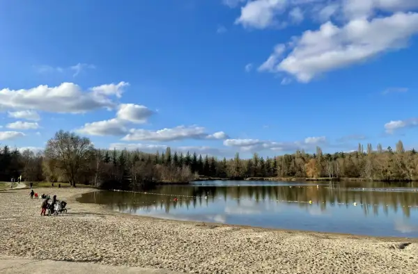 Plage de sable fin au bord d'un lac paisible, avec des familles profitant du soleil. La **cascade** de la **forêt de Fontainebleau** se reflète dans le **lac** sous un ciel bleu parsemé de nuages.