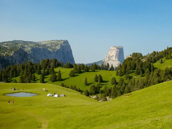 Un campement de tentes et de randonneurs sur une prairie verdoyante, avec des montagnes imposantes en arrière-plan. Le **pas de l'aiguille** est visible au loin.