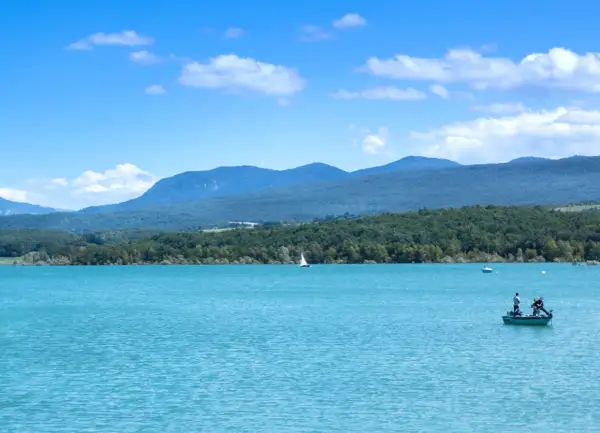 Le lac de Montbel, aux eaux turquoise, accueille un voilier et un petit bateau de pêche sous un ciel bleu parsemé de nuages.